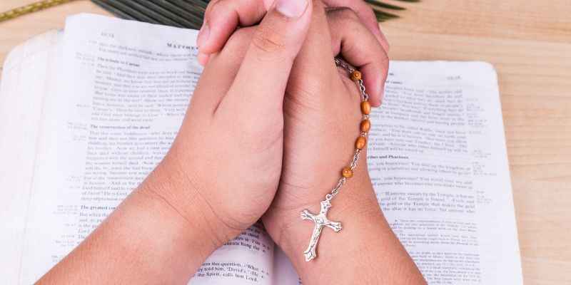 hands closed in prayer holding a rosary over a Bible